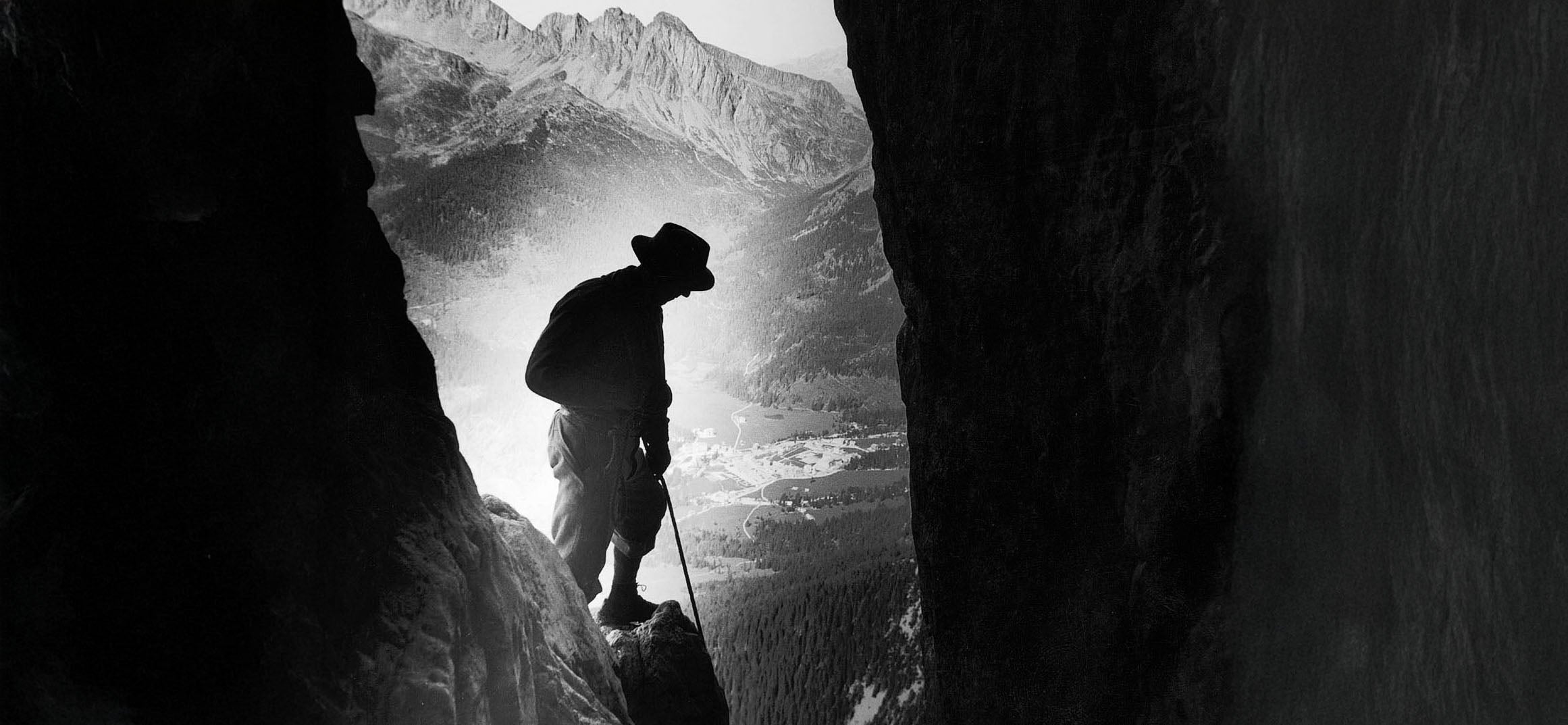 Black and white silhouette of man standing between rocks and mountain ridge in the background