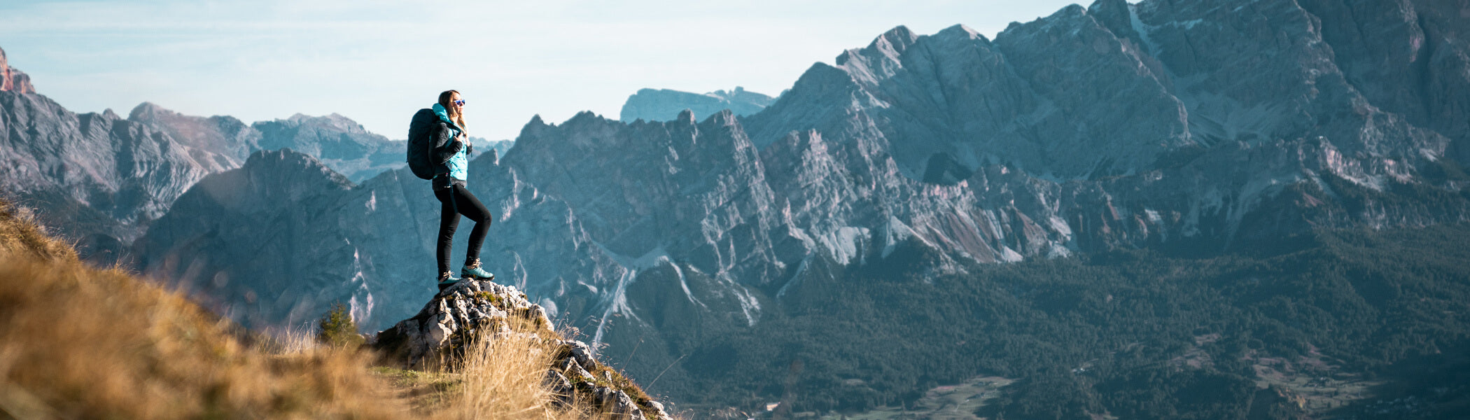 A woman standing on a boulder looking out at the mountain range in front of her. 