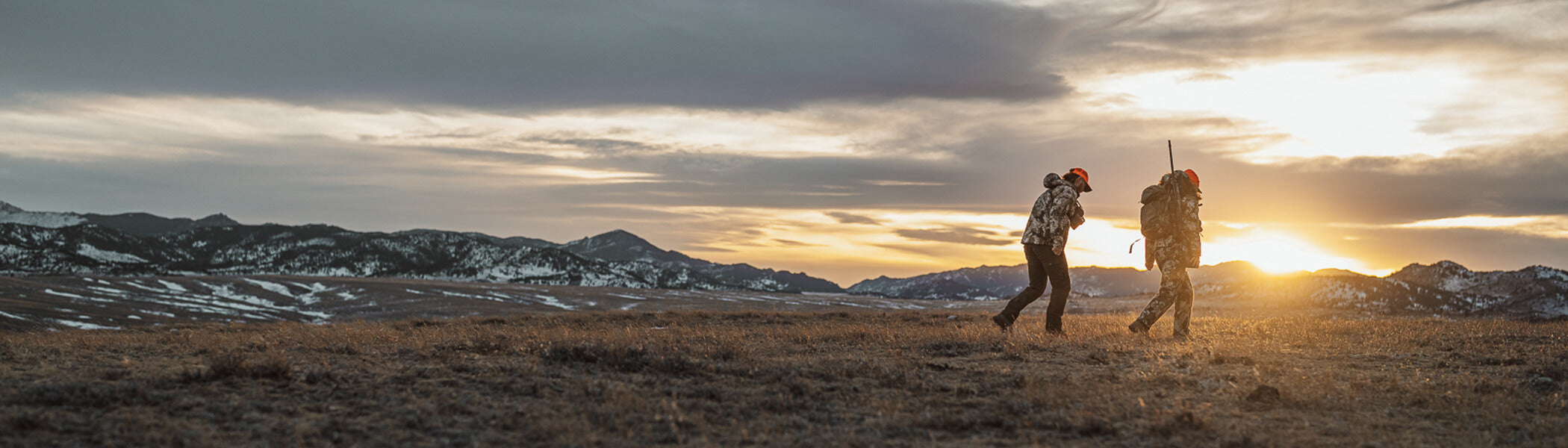Two hunters walking in an open field with mountains in the background. 