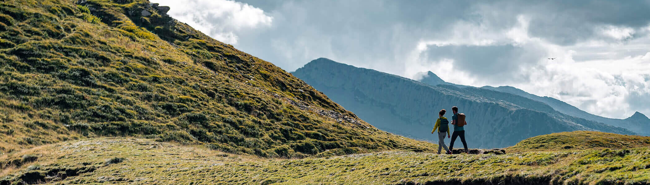 Two hikers walking across a grassy field with mountains in the background. 